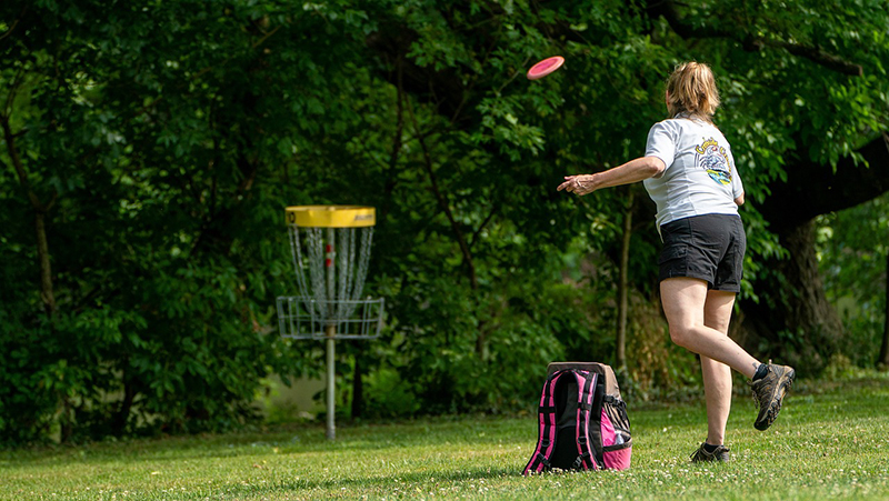 En person ute i naturen kastar en frisbee mot ett discgolfhål. 